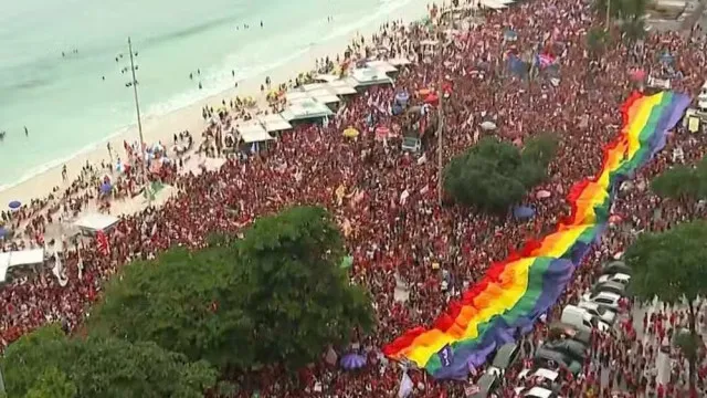 Uma multidão de pessoas em uma avenida larga à beira-mar, segurando faixas e bandeiras, sob um céu claro.