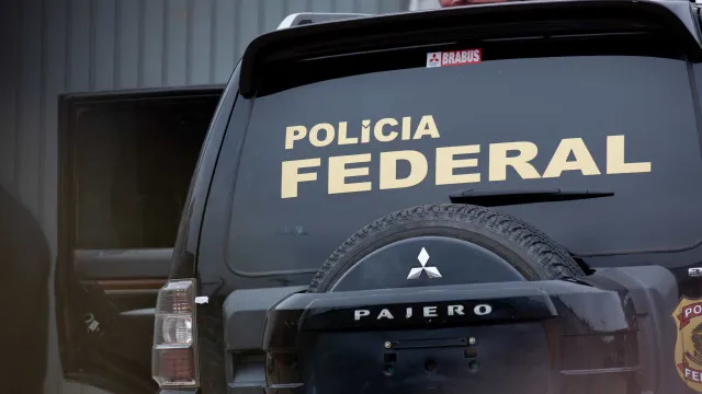 Uma mão segurando um maço de dinheiro ao lado de uma urna, com um distintivo de polícia ao fundo.