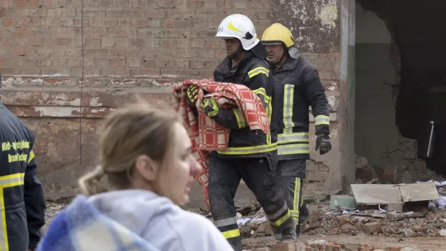 Ônibus destruído em uma rua devastada pela guerra, com fumaça ao fundo e destroços espalhados.