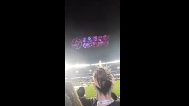 Torcedores em um estádio à noite, com drones formando um display de luz no céu, alguns mostrando um logotipo. O ambiente é de excitação e leve frustração.