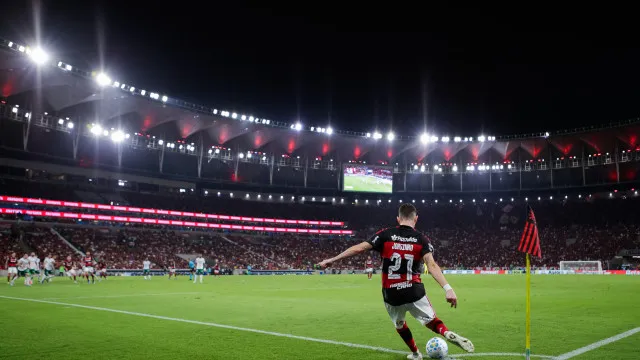 Visão ampla de um estádio de futebol lotado com torcedores, iluminado por luzes fortes sob um céu crepuscular.