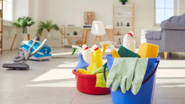 Uma pessoa com luvas de proteção em um ambiente de cozinha, limpando uma superfície com um pano. Há frascos genéricos de produtos de limpeza no balcão, mas sem marcas. A cena transmite uma sensação de limpeza e organização.