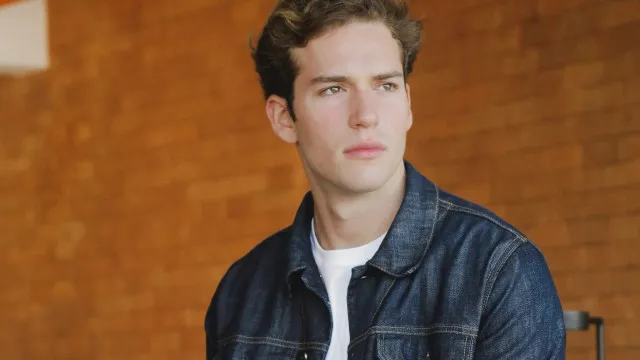 Homem jovem em cenário teatral, sentado em momento de reflexão com iluminação suave ao fundo