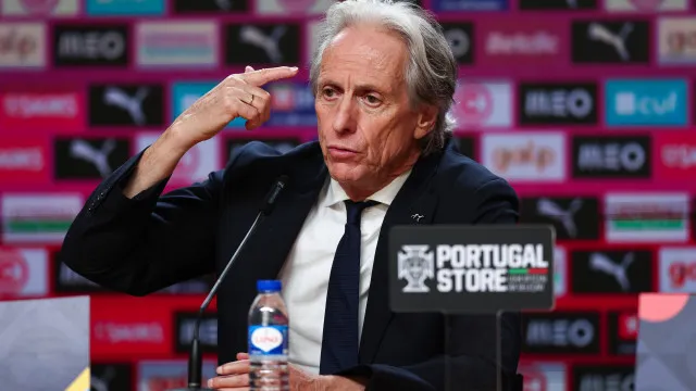 Um treinador de futebol fala em uma coletiva de imprensa, com microfones à frente e um fundo desfocado de estádio.