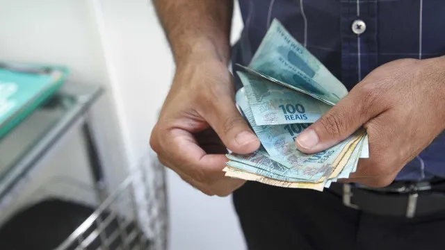 Uma mão contando uma pilha de notas de reais, com algemas e documentos oficiais desfocados ao fundo.