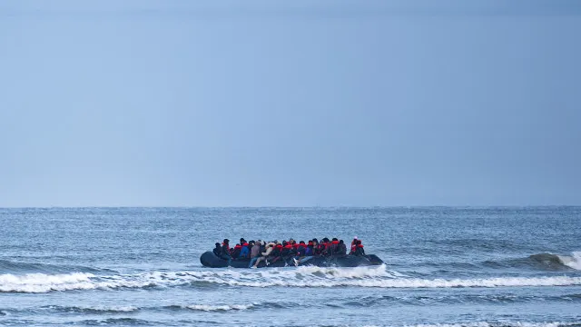Embarcação superlotada em águas agitadas com navio de resgate ao fundo em dia nublado