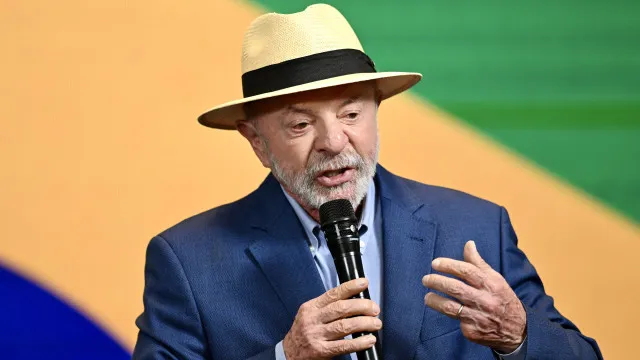 Sala de reunião do governo com mesa comprida, documentos e a bandeira brasileira; pessoas em discussão formal sobre políticas