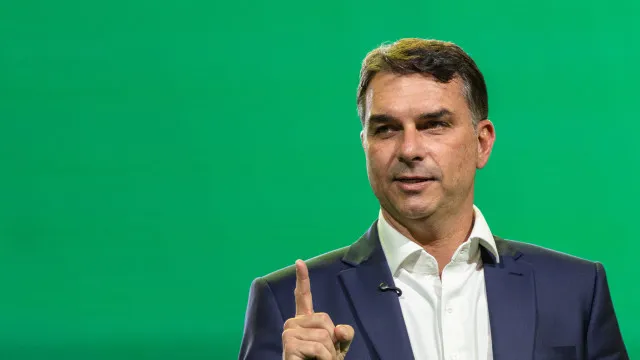 Uma mão segura um smartphone mostrando uma interface genérica de apostas esportivas, com figuras abstratas e embaçadas ao fundo que sugerem um debate político.