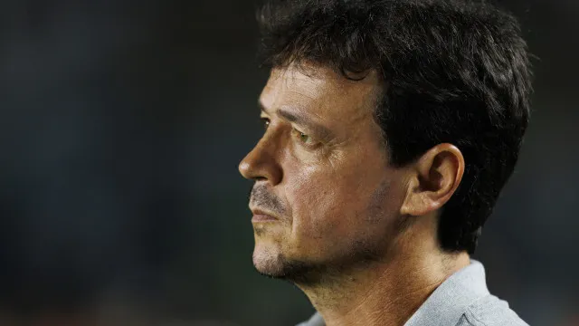 Um treinador de futebol solitário, com semblante abatido, na lateral de um campo, com um estádio desfocado ao fundo.