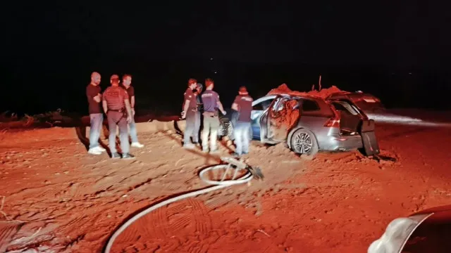 Carcaça carbonizada de um carro em um campo rural ao anoitecer, com fumaça subindo. A cena é desolada.