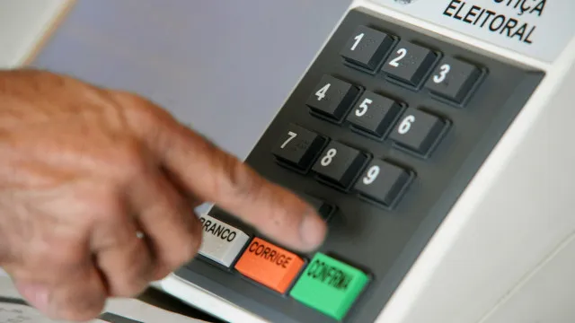 Pessoa em uma mesa cercada por panfletos de campanha e materiais sobre candidatos, pesquisando e anotando informações para decidir seu voto