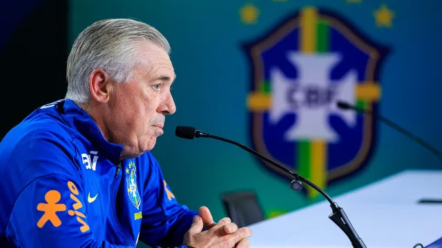 Campo de futebol com jogadores em movimento, estádio ao fundo sob holofotes, bola no ar.